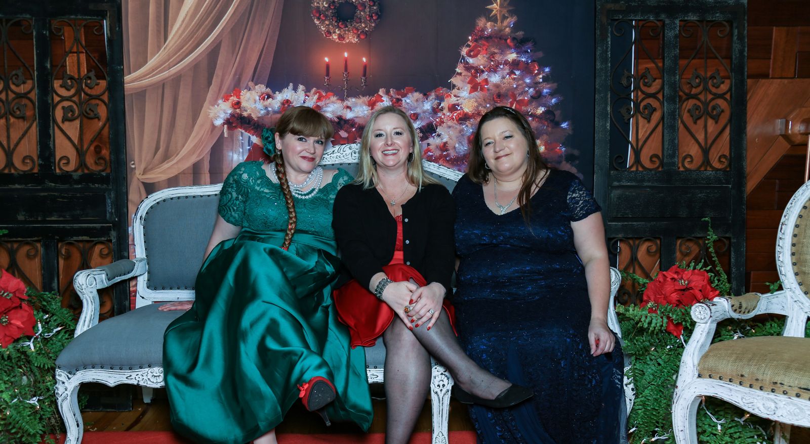 Three of the elves in holiday dresses on a settee in front of a lit Christmas tree and poinsettias