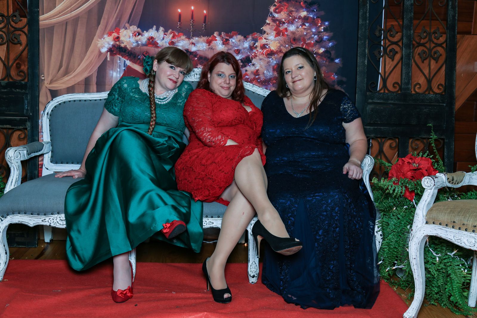 Three of the elves in holiday gowns on a settee in front of a lit Christmas tree, candles and poinsettias